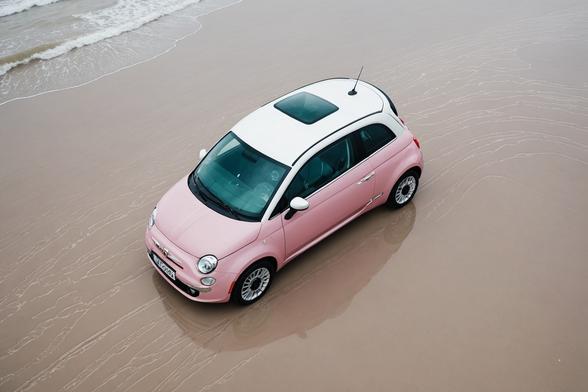 Vue aérienne d'une Fiat 500 couleur rose sur du sable blanc immaculé avec son reflet sur le sable mouillé et un ciel dégagé.