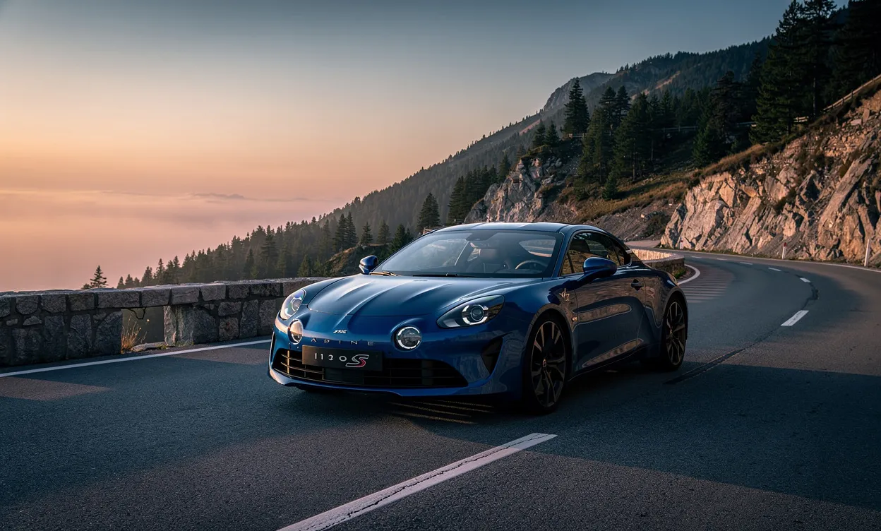 Une Alpine A110 S bleue est garée sur une route de montagne sinueuse au crépuscule, éclairée par une lumière dorée.