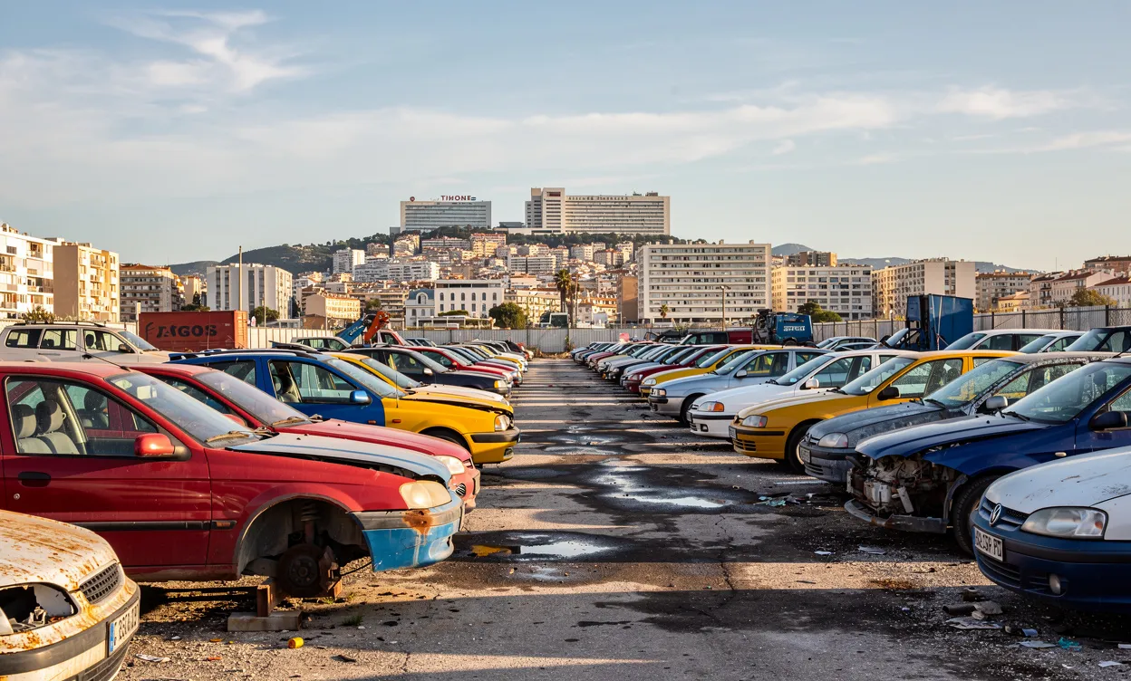 Vue d’une casse automobile colorée sous le soleil méditerranéen à Marseille, avec la silhouette de la ville et l’hôpital de la Timone en arrière-plan.