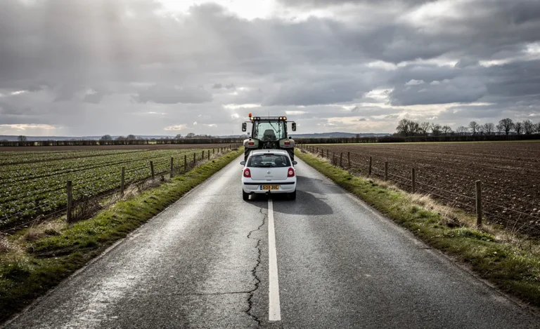 Une petite voiture suit un tracteur lent sur une route rurale étroite bordée de champs sous un ciel nuageux.